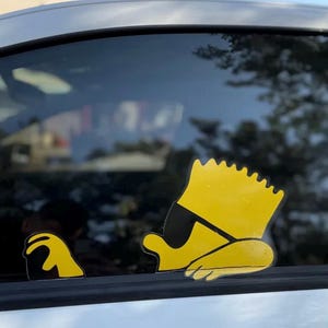 Peut inclure: Un autocollant en vinyle jaune de Bart Simpson portant des lunettes de soleil, appuyé contre une vitre de voiture. L'autocollant est une image de dessin animé du personnage de la série télévisée Les Simpson.