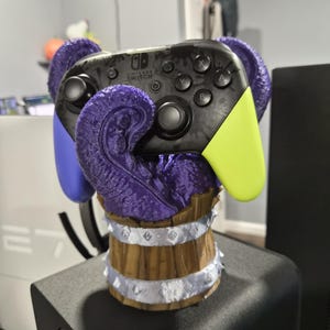 Puede incluir: Un mando Nintendo Switch Pro descansa sobre un soporte personalizado. El mando es negro con detalles en gris y agarres verdes y azules. El soporte presenta un diseño de tentáculo morado y una base de barril de madera con adornos plateados.