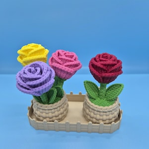 May include: Four crochet roses in a beige basket tray. The roses are in shades of yellow, pink, purple, and red. Each rose has green leaves and is potted in a beige basket. The tray has a decorative fence design.