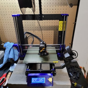 Op de afbeelding: Een blauwe en zwarte 3D-printer met een filamentrol, een bouwplaat en een display dat de tekst "ORIGINAL PRUSA" en de status "Prusa 13 MK3S OK." weergeeft.