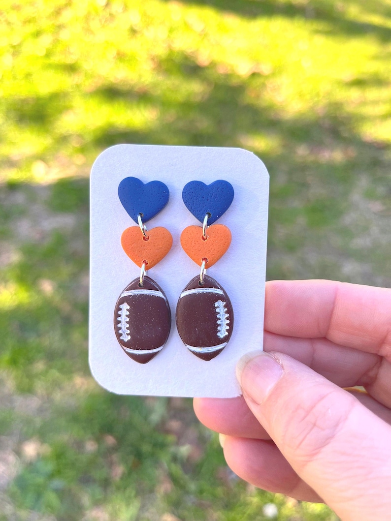 May include: A pair of dangle earrings featuring a blue heart at the top, an orange heart in the middle, and a brown football with white laces at the bottom. The earrings are attached to a white display card with a rounded rectangular shape.