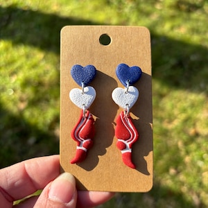 May include: A pair of handmade earrings featuring a patriotic design. Each earring has three heart-shaped elements: a dark blue heart, a white heart, and a red, flame-like shape with white accents. The earrings are displayed on a brown card.