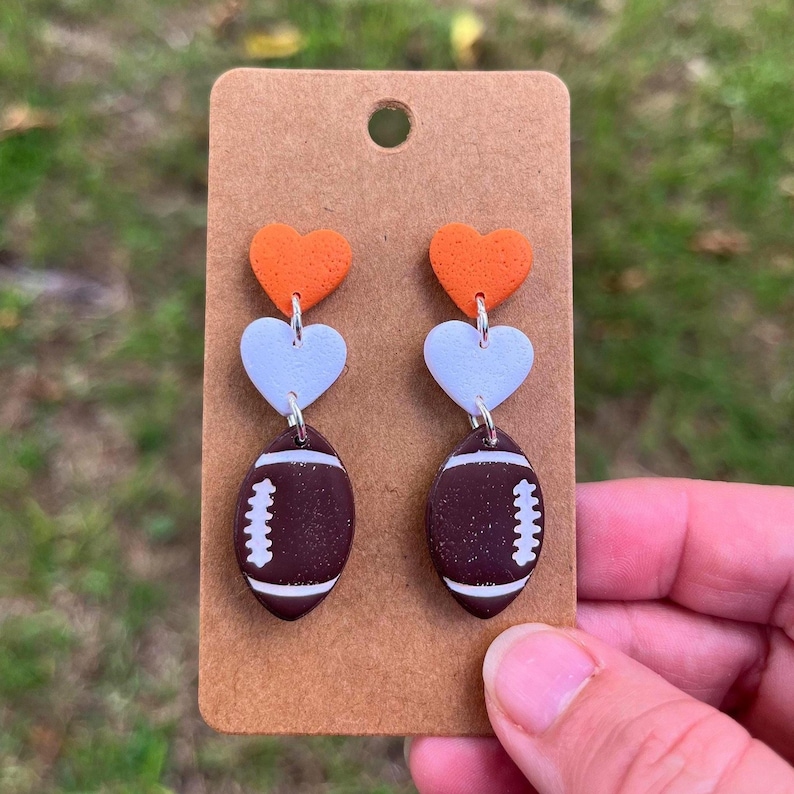 May include: A pair of dangle earrings featuring a vertical arrangement of three charms: a textured orange heart, a white heart, and a brown football with white laces. The earrings are attached to a rectangular brown cardboard display card.