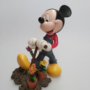 Può includere: Una statuetta di Topolino vestito con una tuta, che tiene uno strumento da giardinaggio e sta in piedi in un pezzo di terra con dei fiori.