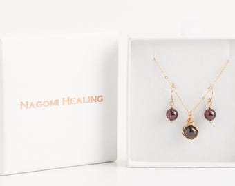 May include: A set of garnet jewelry in a white box. The set includes a necklace and earrings with a gold chain and a dark red garnet stone pendant. The box has the text "NAGOMI HEALING" printed on it.