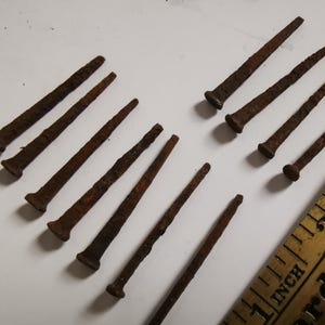 May include: A selection of old, rusty nails with flat heads and pointed ends. The nails are of varying lengths and are laid out on a white surface. A wooden ruler with inch markings is also visible.