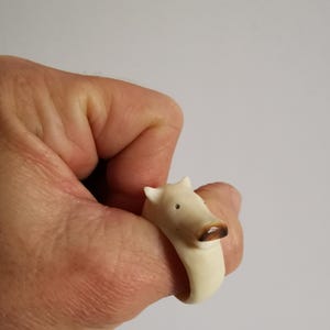 May include: A cream-coloured ring featuring a pig design. The pig's snout is brown, and it has small black eyes. The ring is worn on a finger, showcasing the animal's head as a decorative element. The ring is made of a smooth material.