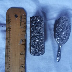 May include: A silver-toned hand mirror and a small silver-toned brush with floral designs. The mirror is oval shaped and the brush is rectangular. Both items are approximately 10 cm long.