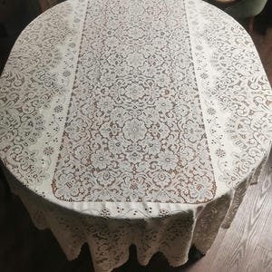 May include: A white oval lace tablecloth with a delicate floral pattern. The tablecloth has a scalloped edge and is ideal for a formal dining setting.