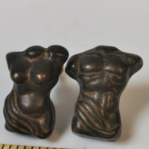May include: Two bronze-coloured torso sculptures, one female and one male, are displayed. The female torso has defined breasts and a draped cloth. The male torso shows muscular definition. The sculptures are on a white surface.