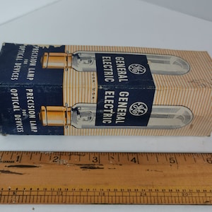 Peut inclure: Une boîte de deux lampes de précision General Electric pour appareils optiques. La boîte est bleue et blanche avec des rayures dorées. Le texte sur la boîte indique "General Electric" et "Lampe de précision pour appareils optiques".