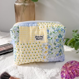 May include: A patchwork cosmetic bag with a floral and gingham pattern, featuring the text "Patchy Berry." A makeup brush, eyeshadow palette, and a purple hair claw are also visible.