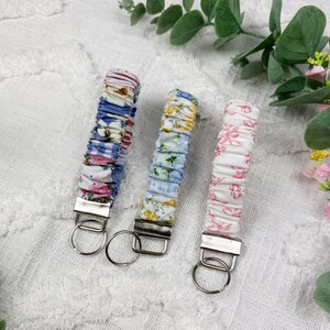 Op de afbeelding: Drie stoffen polssleutelhangers met zilverkleurige metalen hardware en ronde sleutelringen. De linker sleutelhanger heeft een blauw en roze bloemenpatroon, de middelste een blauwe en gele bloemenprint en de rechter een wit en roze bloemendesign.