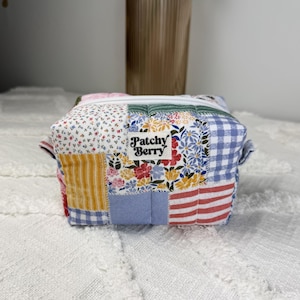 May include: A rectangular quilted pouch featuring a patchwork design of various floral, striped, and gingham fabric patterns in red, yellow, blue, and white. A white zipper runs across the top, and a white fabric label reads Patchy Berry.