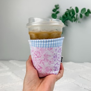Könnte beinhalten: Eine Tassenmanschette aus Stoff mit einem hellblau-weißen Vichy-Karo-Rand oben und einem Patchwork-Körper mit rosa Rosen- und Schmetterlingsmustern sowie einem violetten Blumenmuster. Ein kleines weißes Etikett mit der Aufschrift Patchy ist seitlich.
