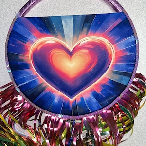 May include: A round, heart-shaped bag with a purple handle and a vibrant heart design. The heart is a gradient of red, orange, and blue, radiating light. The bag is trimmed with sequins and colorful metallic fringe.