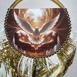 May include: A round decorative piece featuring a dove with outstretched wings, set against a background of hands. The image is framed with gold beads and a gold handle. Gold and white ribbons and tinsel add to the decorative design.