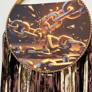 May include: A circular wall hanging featuring a printed image of heavy metal chains breaking apart with golden light. The circular frame is wrapped in gold ribbon and adorned with small metal studs. Long, metallic black and gold fringe hangs below.