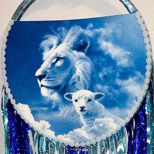 May include: A circular wall hanging featuring a blue and white print of a lion and a lamb surrounded by clouds. The circular frame is bordered by white lace trim and includes long, shimmering blue and silver metallic fringe hanging below.