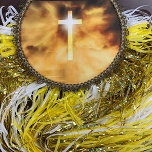 Op de afbeelding: Een religieuze wanddecoratie met een centraal beeld van een wit kruis tegen een gouden hemel. De cirkelvormige afbeelding is omlijst met een kralenrand en omgeven door stromende linten in geel, wit en goud.