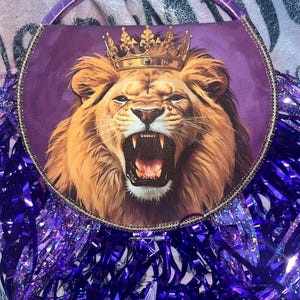 May include: A purple handbag with a roaring lion graphic wearing a gold crown. The lion is set against a purple background and surrounded by purple fringe and a purple handle. The bag has a gold trim.