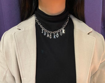 May include: A silver chain necklace with the word "JUNG KOOK" spelled out in silver charms hanging from it. The necklace is worn by a person wearing a black mock neck shirt and a beige blazer.