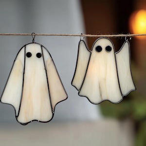 Op de afbeelding: Twee glazen geestenornamenten hangen aan een touwtje. De geesten hebben een crèmewitte body met zwarte ogen en zijn zwart omlijnd. De geest links heeft een afgeronde vorm, terwijl die rechts de armen uitstrekt.