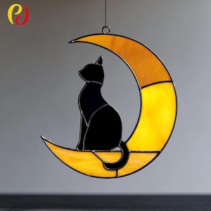 May include: A stained glass suncatcher featuring a black cat silhouette perched on a yellow crescent moon. The moon has a textured, stained glass appearance with a black border. The suncatcher is suspended by a black chain.