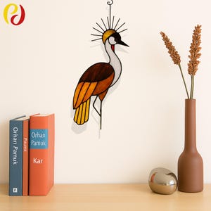 May include: A stained glass crane wall hanging with a crown-like design. The bird features brown, orange, and yellow hues, with a white neck and a black beak. The artwork is suspended by a chain. A brown vase with dried flowers and two books are on a wooden surface.