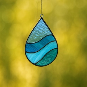 May include: A stained glass teardrop suncatcher with a black metal frame. The design features layers of blue and teal glass, resembling ocean waves. A small chain is attached for hanging. The background is a blurred yellow-green.