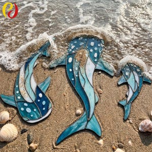 May include: Three stained glass whale shark figures in varying sizes, crafted with blue, teal, and white glass pieces joined by dark metal solder lines. Each figure features a spotted pattern and is positioned on wet sand near the ocean surf.
