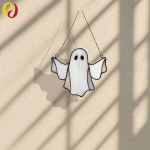 May include: A white stained glass ghost with outstretched arms, hanging from two thin wires. The ghost has two black oval eyes and a black outline. The image is set against a beige background with shadows.