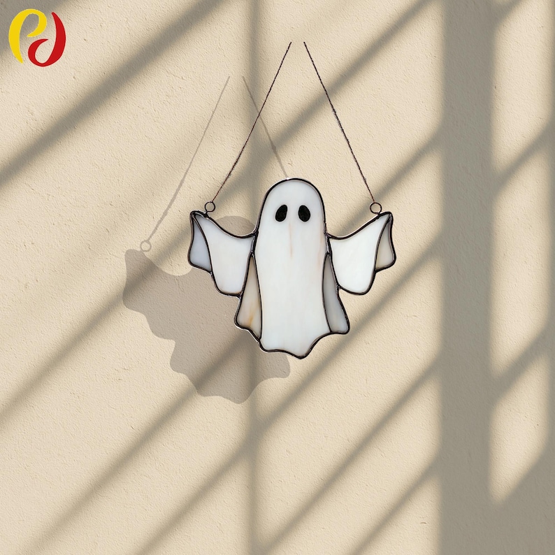 May include: A white stained glass ghost with outstretched arms, hanging from two thin wires. The ghost has two black oval eyes and a black outline. The image is set against a beige background with shadows.