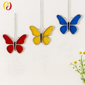 May include: Three stained glass butterfly ornaments in red, yellow, and blue, each with a black body and silver chain for hanging. The butterflies are suspended against a white background.