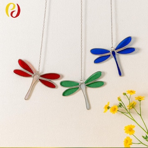 May include: Three stained glass dragonfly ornaments in red, green, and blue hang from silver chains. The dragonfly bodies are silver, and the wings are vibrant colors. Yellow flowers are in the lower right corner.