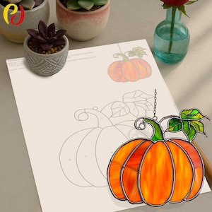 May include: A stained glass pumpkin in shades of orange and yellow, with green leaves and a metal chain. The pumpkin is displayed against a light-coloured paper with a pumpkin outline. A small succulent plant in a decorative pot is also visible.