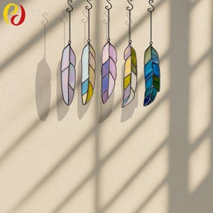 May include: Five stained glass feather ornaments in various colors hang from black chains. The feathers are in shades of purple, blue, yellow, and green, with a black outline. The ornaments are suspended against a neutral wall.