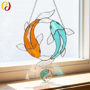 Peut inclure: Suncatcher en vitrail avec deux poissons koi, un orange et un turquoise, formant un cercle. Un symbole yin-yang est suspendu en dessous. Le capteur solaire est suspendu par une chaîne argentée.
