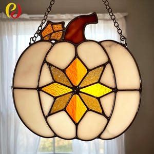 Peut inclure: Décoration de citrouille en vitrail avec un corps blanc, un centre en étoile orange et jaune, et une tige et une feuille marron. La citrouille est suspendue par une chaîne, idéale pour la décoration d'automne.