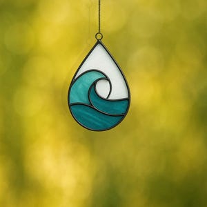 May include: Teardrop-shaped stained glass suncatcher with a teal wave design against a white background. The wave is outlined in black, with a chain for hanging. The background is a blurred yellow and green.