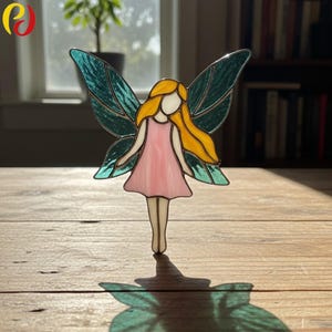 Può includere: Una statuina a forma di fata in vetro colorato con un vestito rosa, capelli gialli e ali turchesi. La fata è in piedi su una superficie di legno, con la sua ombra visibile sotto. Le ali hanno un aspetto strutturato e la figura è delineata in un colore scuro.