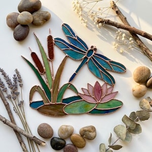 May include: A stained glass suncatcher featuring a blue dragonfly hovering over a pink water lily and brown cattails. The piece is constructed with copper foil solder lines, showcasing translucent glass in shades of teal, blue, green, brown, and pink.