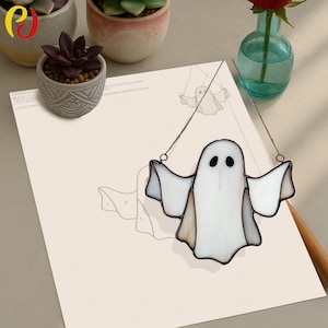 May include: A white stained glass ghost with black eyes and outstretched arms, hanging from a thin wire. The ghost is set against a light-colored background with a drawing of a ghost. The image also includes small potted plants and a vase with a red flower.