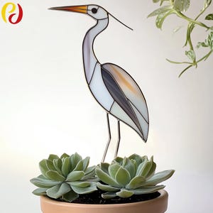 May include: A stained glass heron sculpture in white, grey, and orange. The bird stands on thin metal legs, above two green succulents in a terracotta pot. The heron has a long orange beak. The pot is filled with soil.