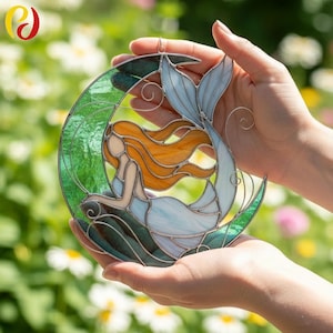 Könnte beinhalten: Buntglas-Suncatcher mit einer Meerjungfrau in einem Halbmond. Die Meerjungfrau hat orangefarbenes Haar und einen hellblauen Schwanz. Der Mond ist grün und der Körper der Meerjungfrau ist hellblau. Der Suncatcher wird vor einem verschwommenen grünen Hintergrund gehalten.