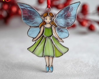May include: A stained glass fairy ornament with a red ribbon for hanging. The fairy has a green dress, blue wings, and brown hair. The ornament is made with a silver metal frame.