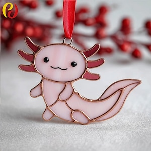 May include: A pink axolotl ornament with a copper-colored outline and a red ribbon for hanging. The axolotl has a smiling face with black eyes and a wavy mouth. The ornament is made of glass and is a decorative item.