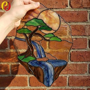 May include: A stained glass suncatcher featuring a landscape scene with a brown tree, green foliage, a blue waterfall flowing into a pool, and a large amber sun in the background. The piece is constructed with dark metal solder lines.