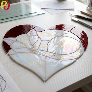 Puede incluir: Un atrapasoles de vidrieras en forma de corazón con el diseño de un gato durmiendo. El gato está hecho de piezas de vidrio blanco iridiscente, mientras que el borde del corazón es de vidrio rojo oscuro, unidos por finas líneas de soldadura.