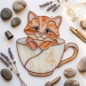 May include: A stained glass suncatcher featuring an orange tabby cat sitting inside a white teacup. The cat has detailed glass segments with copper foil edges, dark eyes, and paws resting on the rim of the cup. The piece includes a small metal loop.